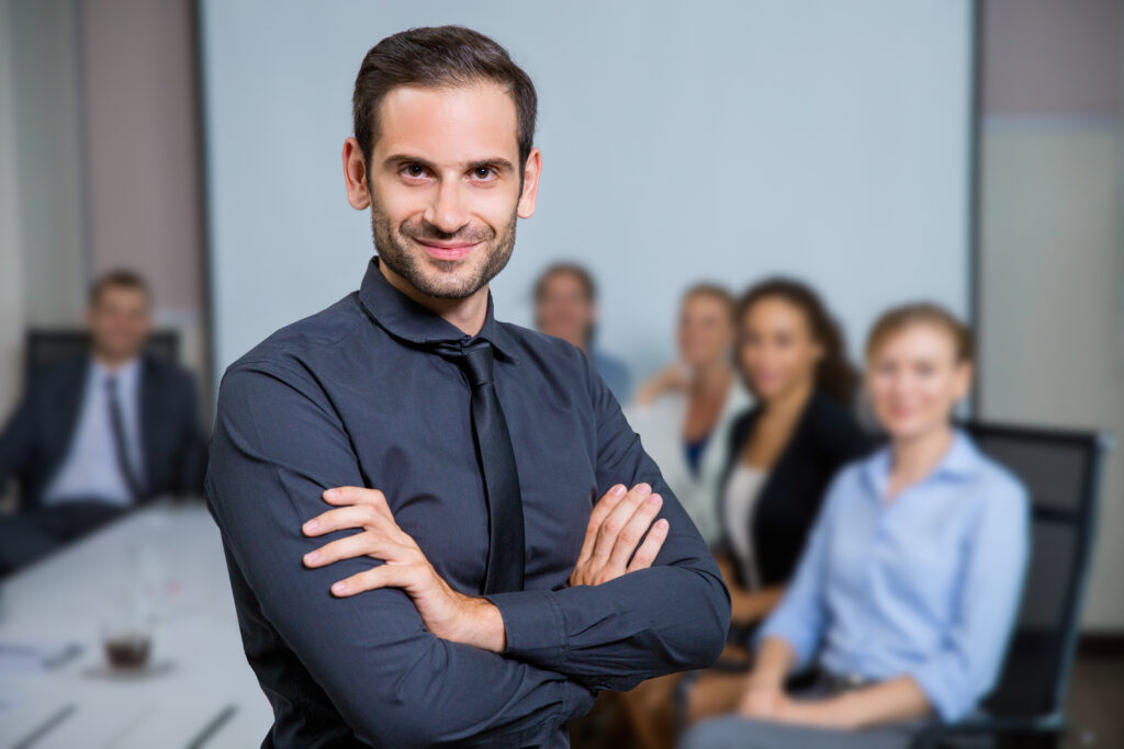 Portrait of successful business leader standing with crossed arms and smiling, business people sitting at office desk on background