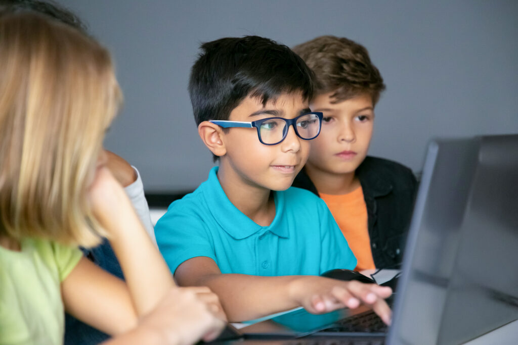 Asian boy typing on laptop keyboard and classmates sitting at table, watching him and doing task together. Pupils using computers during study. Childhood, communication and digital education concept